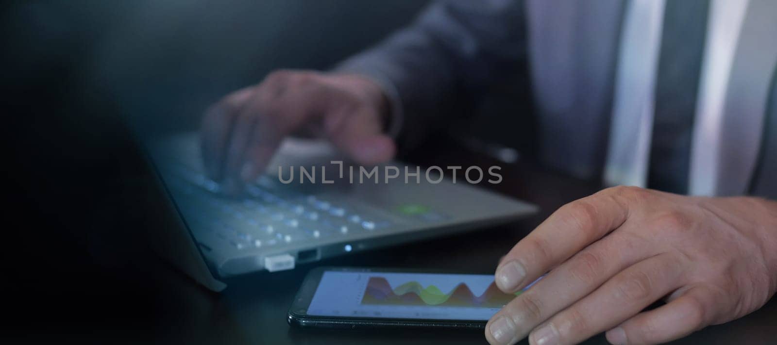 Hands of an unrecognizable man in a suit working with a computer and a smartphone.