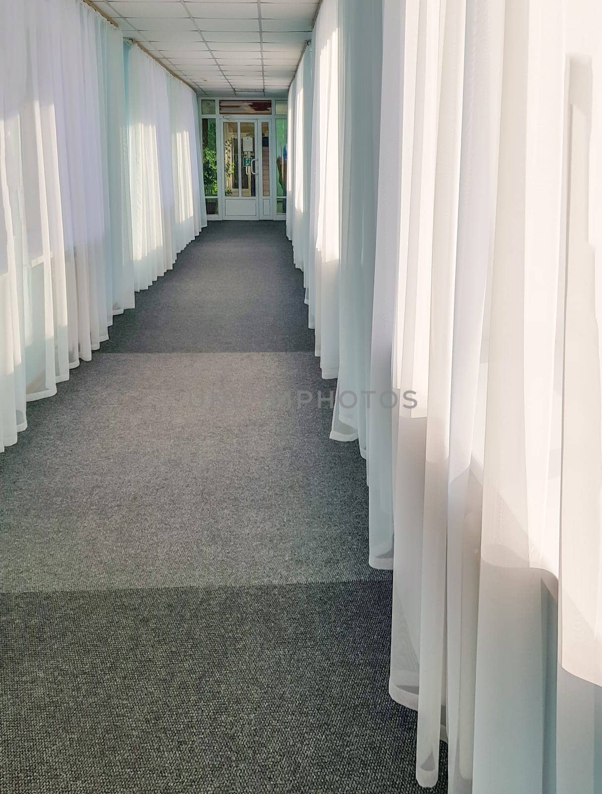 The prospect of a long hotel corridor with transparent curtains on large windows and glass doors.