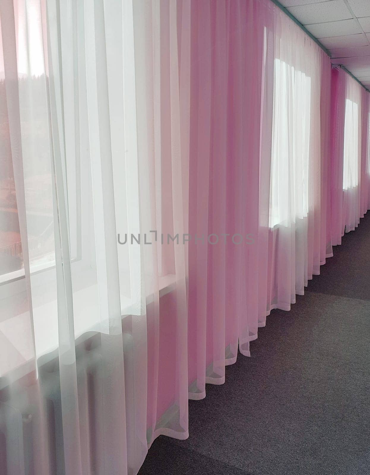 The prospect of a long hotel corridor with transparent curtains on large windows and pink tinting.