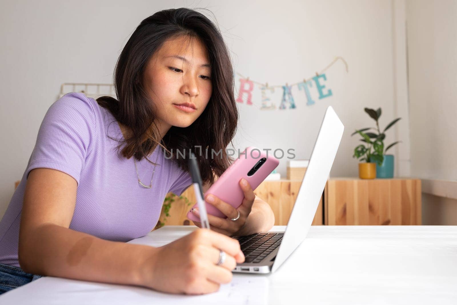 Teenage female college student doing university paper, researching with laptop and mobile phone handwriting information on piece of paper. Education concept.