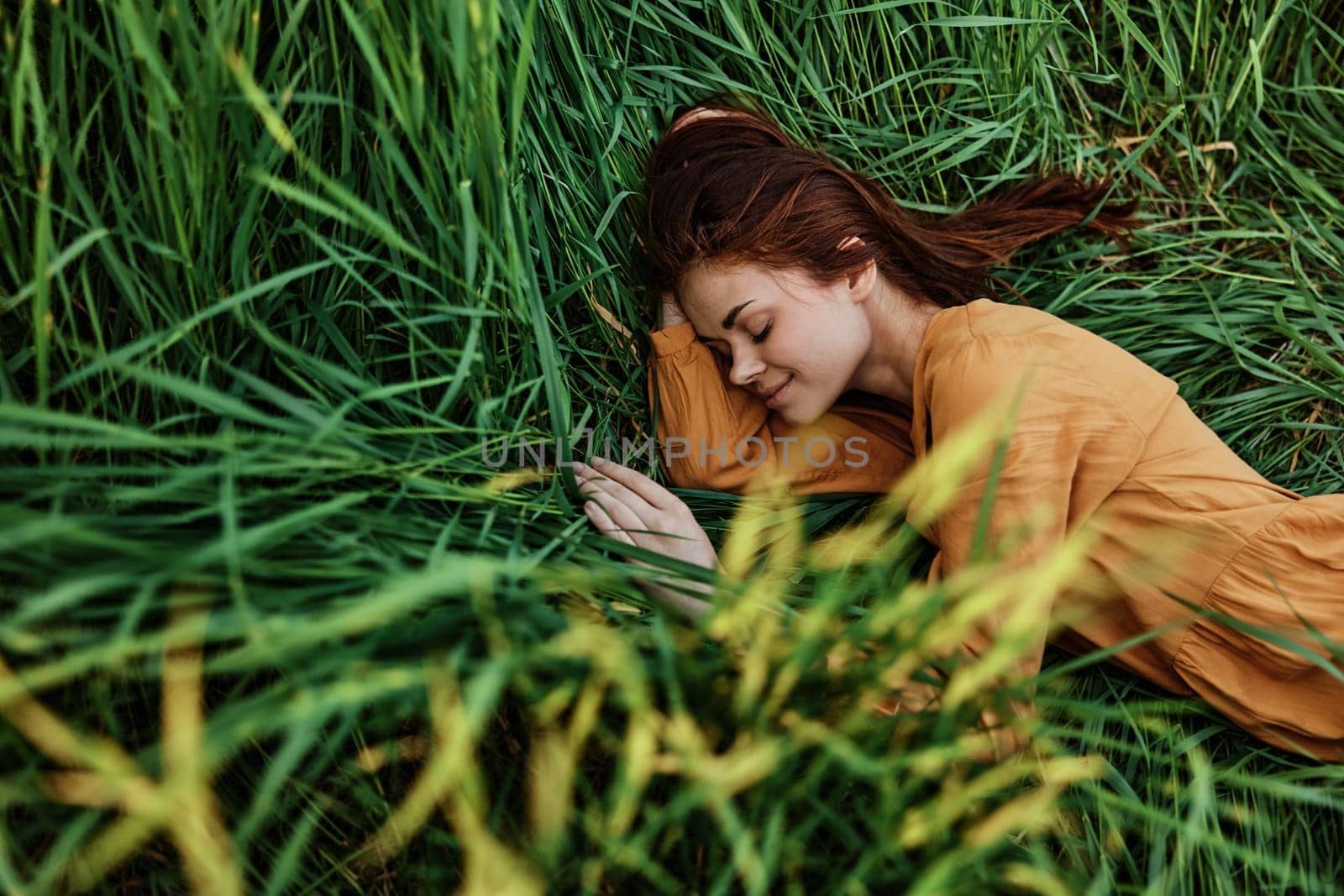 a close horizontal photo of a pleasant woman in a long orange dress resting lying in the tall grass with her eyes closed in sunny weather at sunset with her arms outstretched. Street photography, the theme of privacy with nature. High quality photo