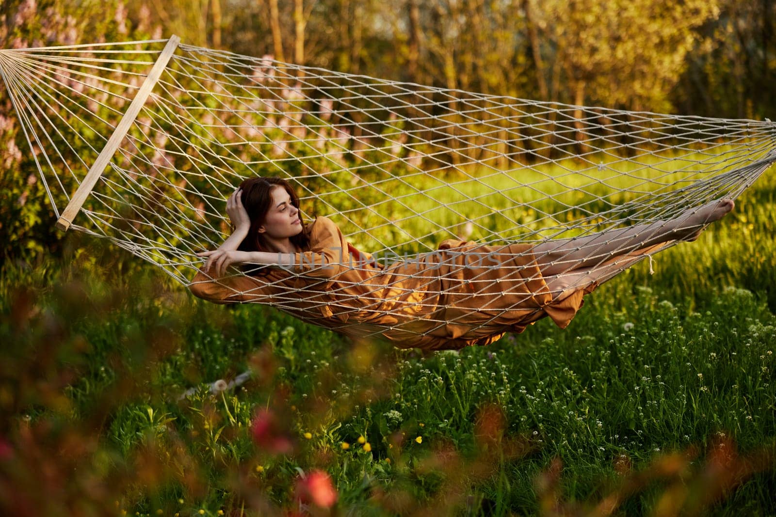 a beautiful woman is resting in nature lying in a mesh hammock in a long orange dress looking to the side, propping her head with her hand. Horizontal photo on the theme of recreation by Vichizh