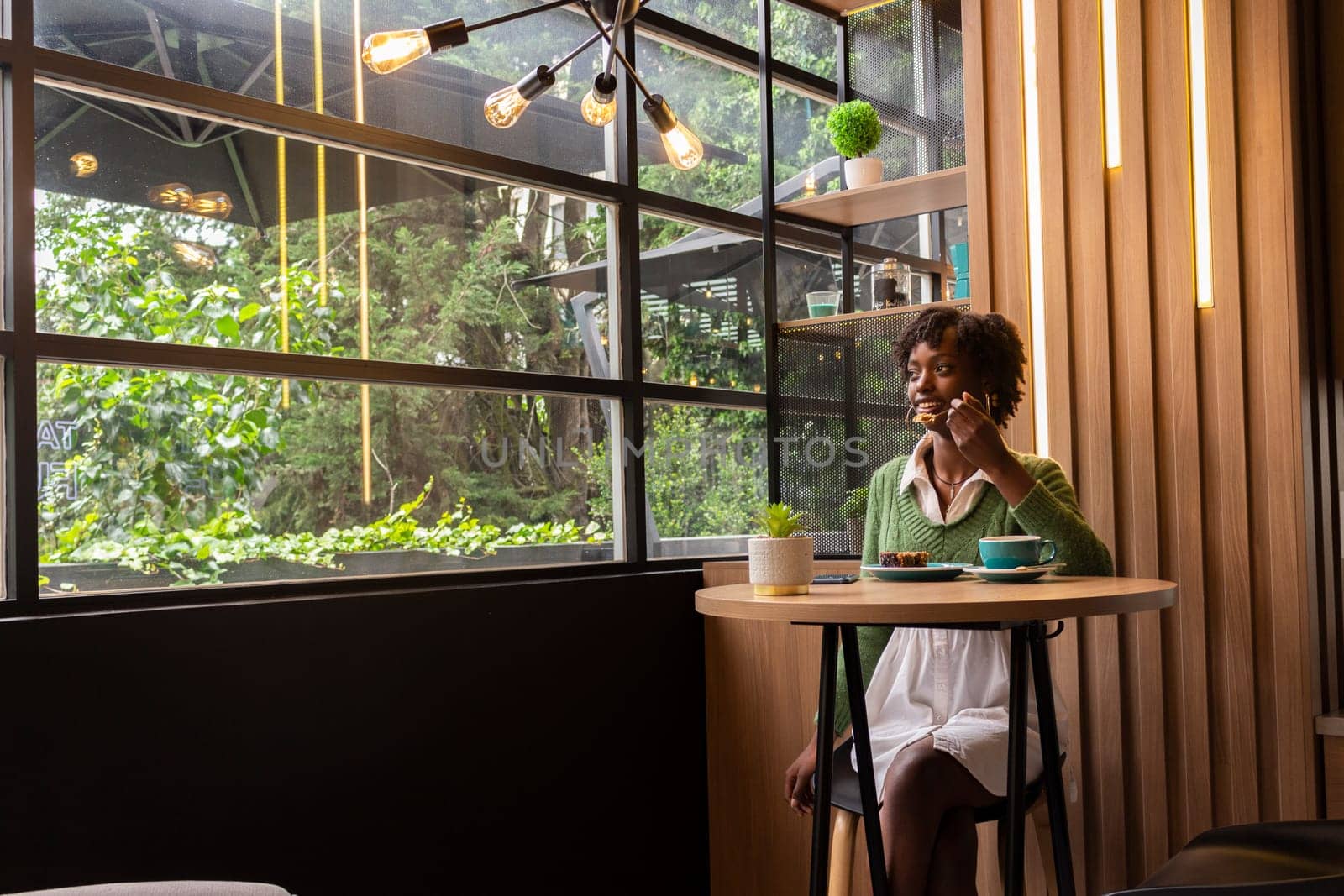 copyspace of african american girl having a cake and a coffee sitting in a cafe looking happily through a window at the city. High quality photo