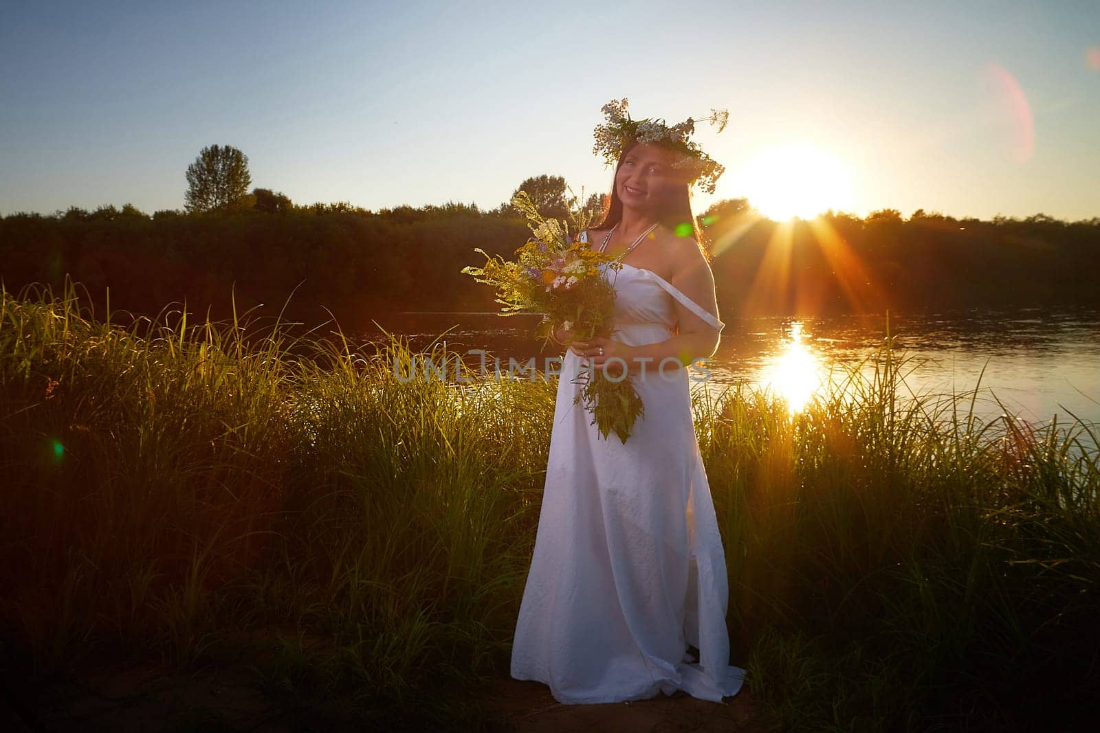 Adult mature brunette woman in a white dress, sundress and a wreath of flowers in summer by the water of river or lake in evening at sunset. Celebration of the Slavic pagan holiday of Ivan Kupala