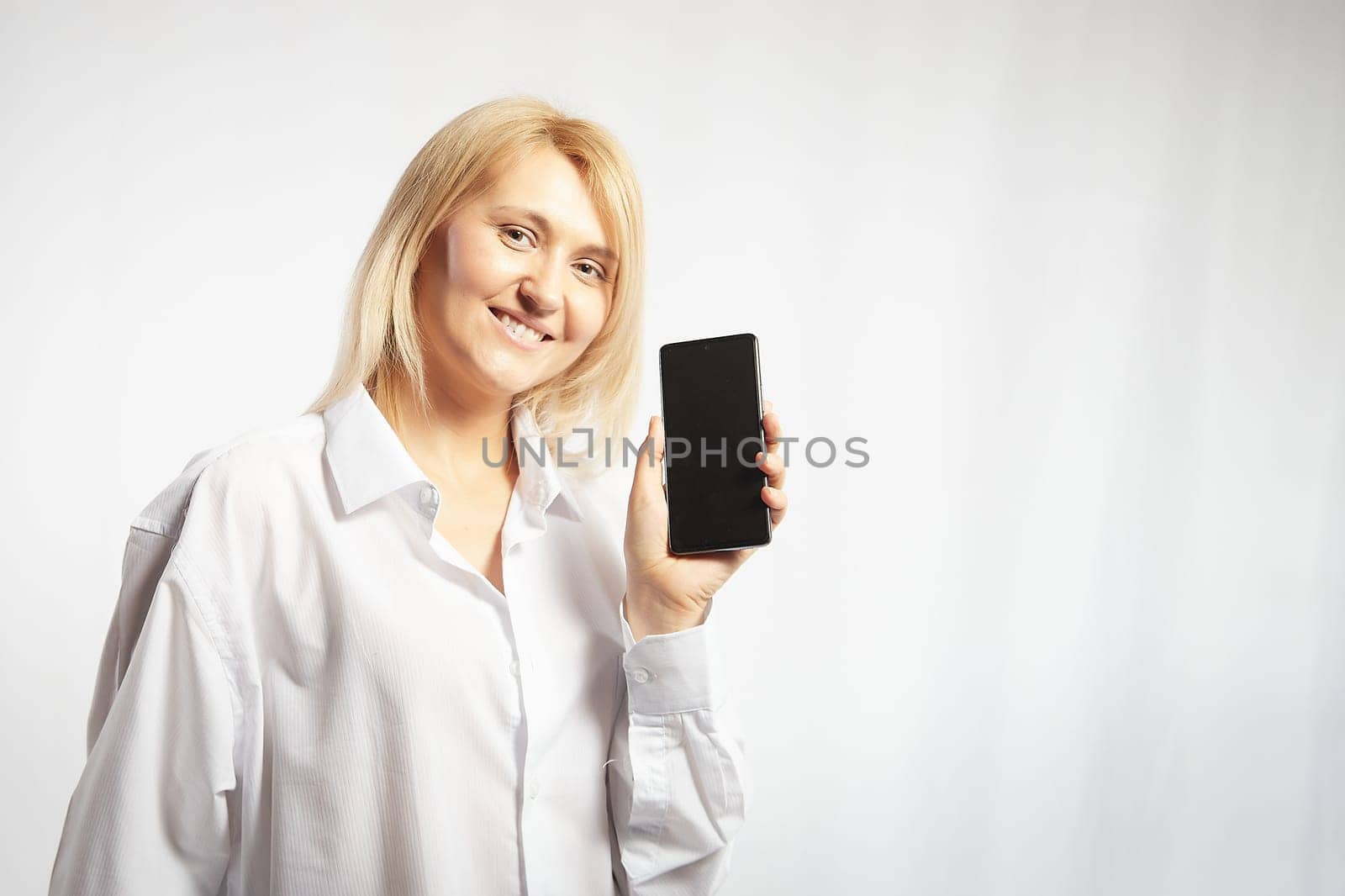 Portrait of a smiling casual blonde woman in shirt holding smartphone over white background. The girl is chatting and taking selfies. Business lady with a cell phone. Copy space by keleny