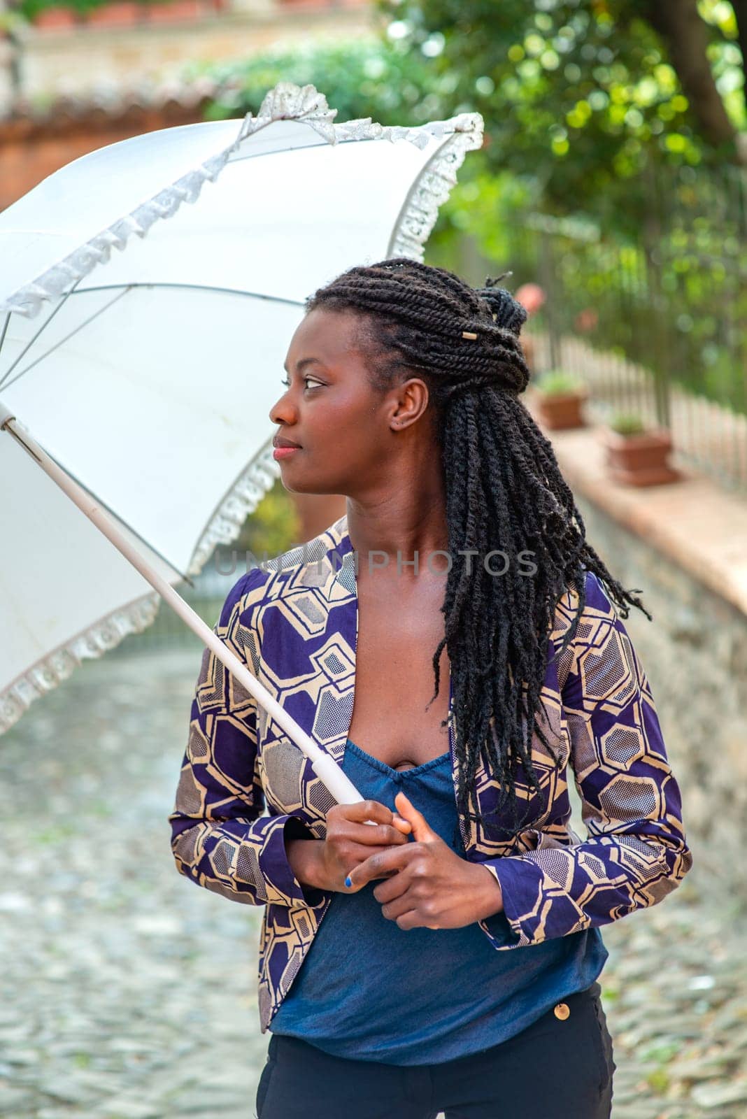 Portrait of an African woman with dreadlocks braid with umbrella parasol. Happy young woman feeling confident in her style. Fashionable woman standing in the street of old village against stone wall.