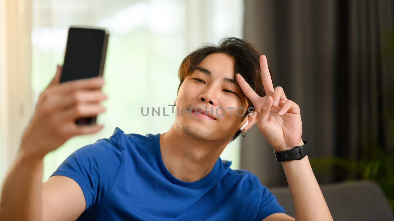 Smiling young asian taking selfie on smartphone and showing V gesture with hand.