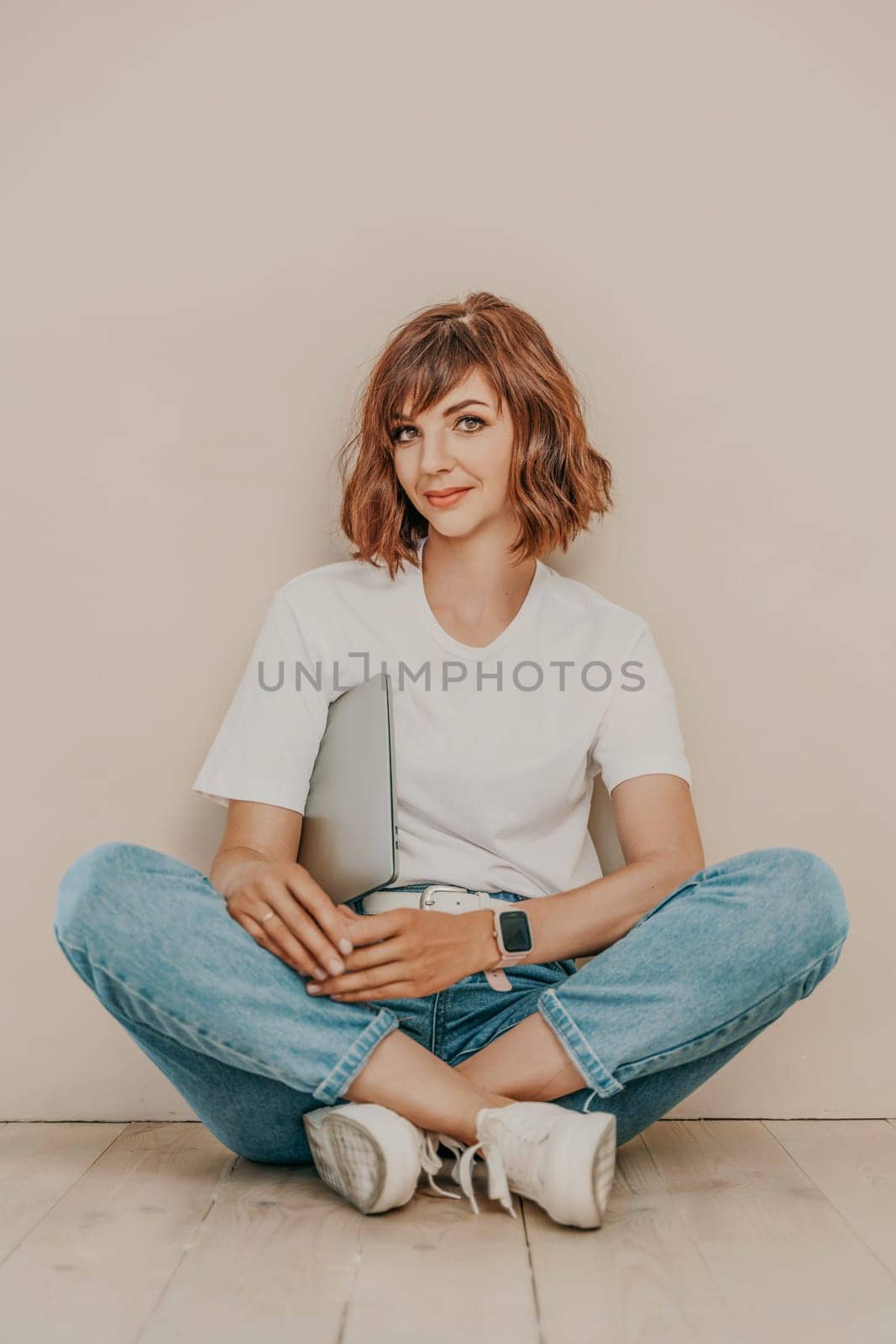 A brunette sits on the floor with a laptop on a beige wall background. She is wearing a white T-shirt, jeans and white sneakers. by Matiunina