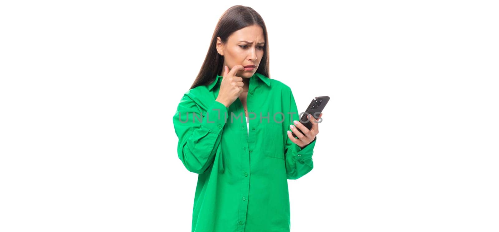 young caucasian brunette lady with makeup dressed in a green shirt and jeans is typing a message on the phone on a white background with copy space.