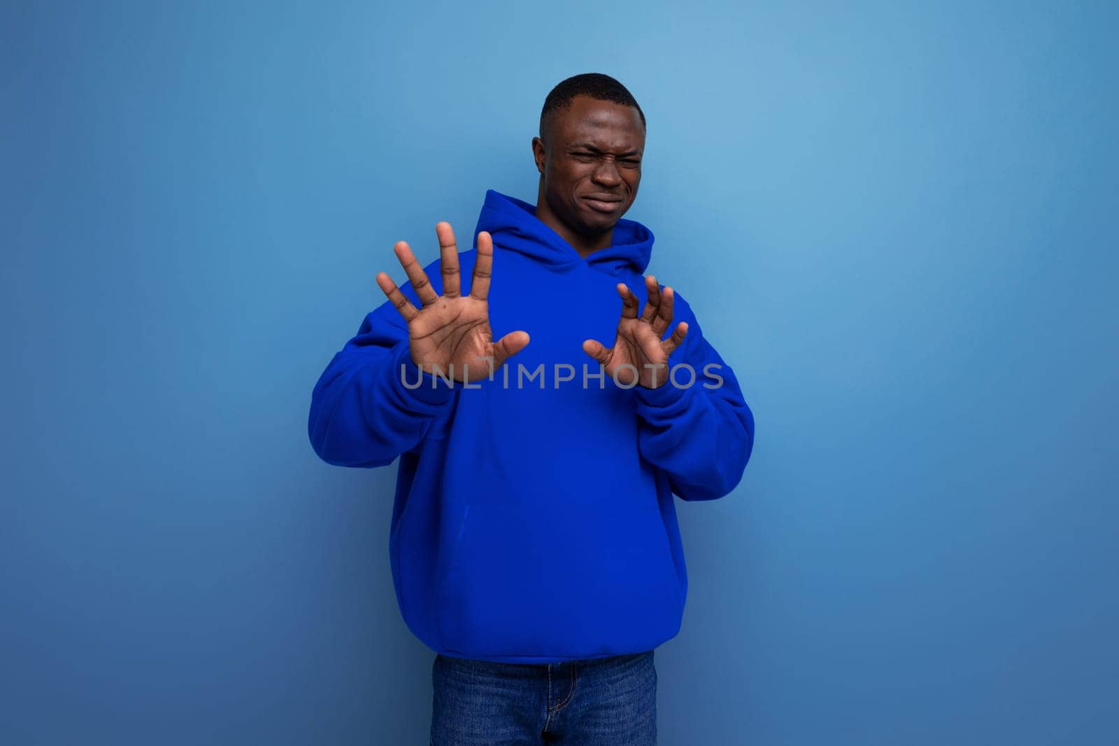 close-up of an emotional african young guy in a blue sweatshirt with a hood on a studio background with copy space by TRMK
