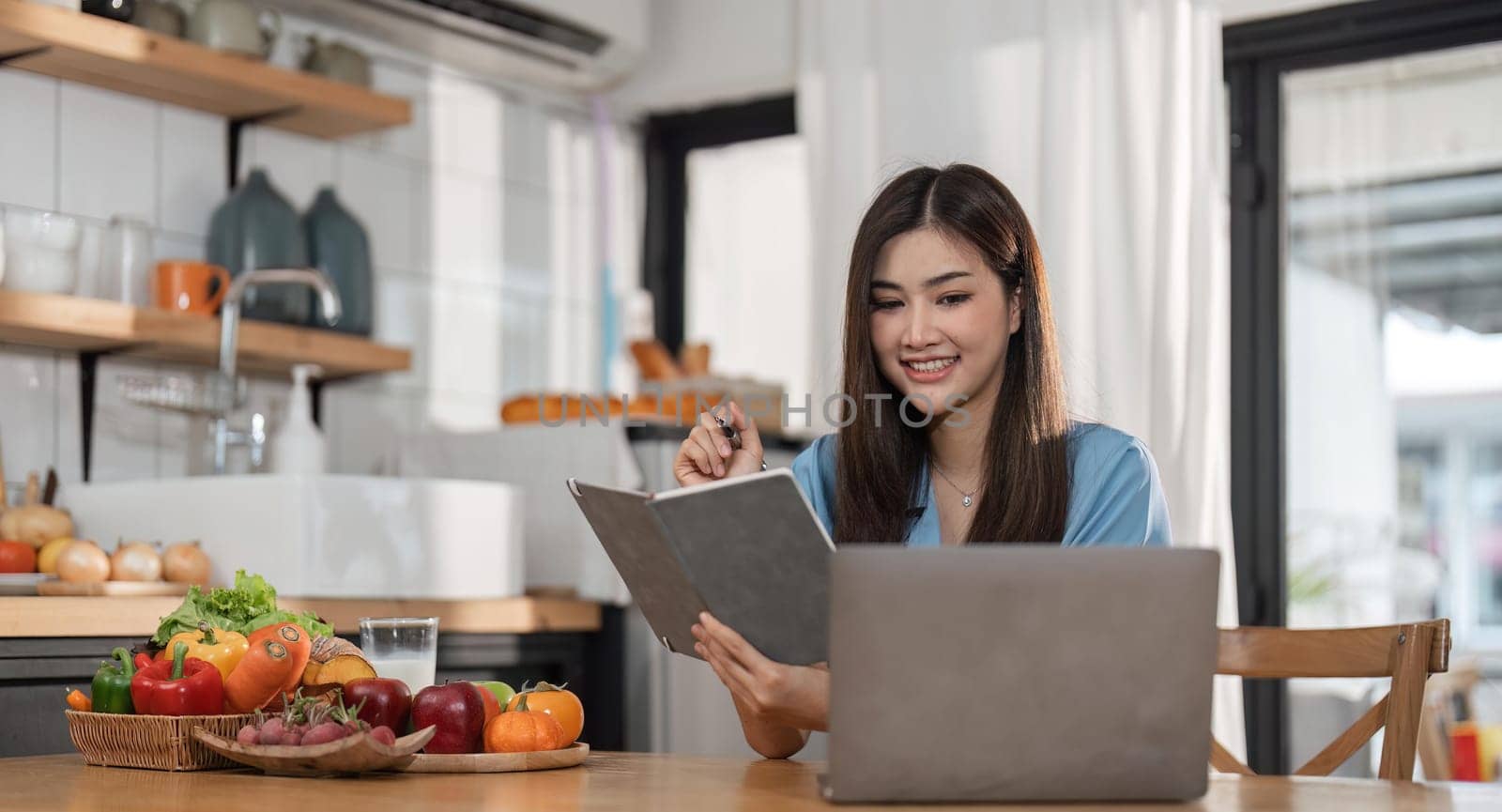 young woman is using laptop computer for remote work or studying online, she takes notes watching webinars or classes at kitchen table at home.