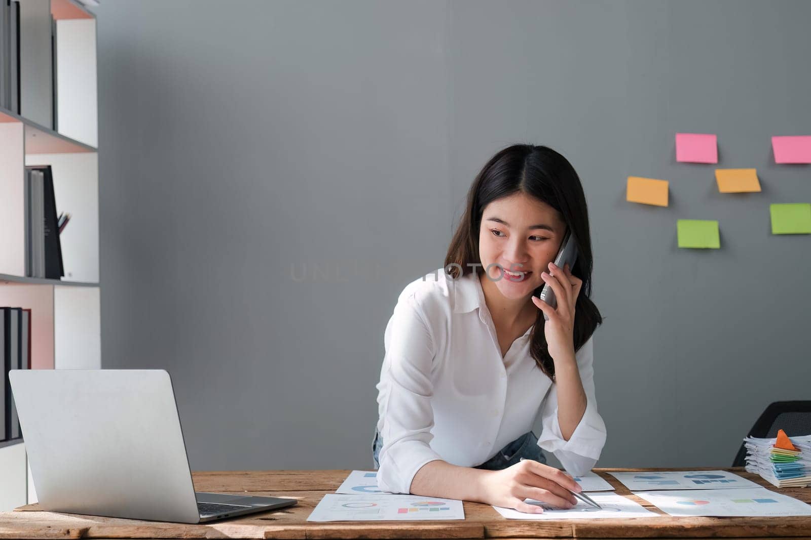 Happy young asian woman talking on the mobile phone and smiling while sitting at her working place in office.