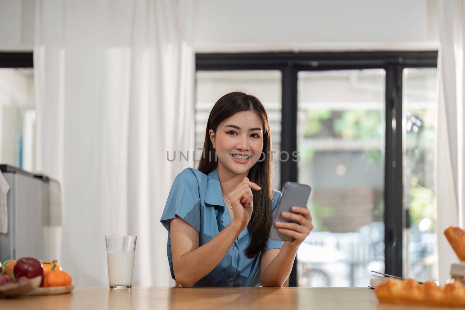 Young woman use cellphone sit at table in kitchen share text messages in social media, enjoy new , watch internet content take break from work on laptop. mobile app usage concept by nateemee