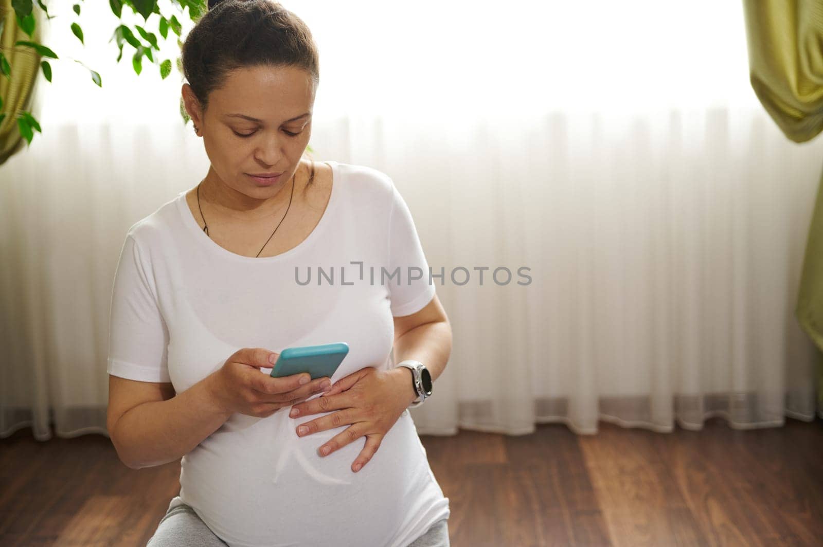 African American beautiful adult woman touching her pregnant belly, using mobile phone during pregnancy yoga practice by artgf