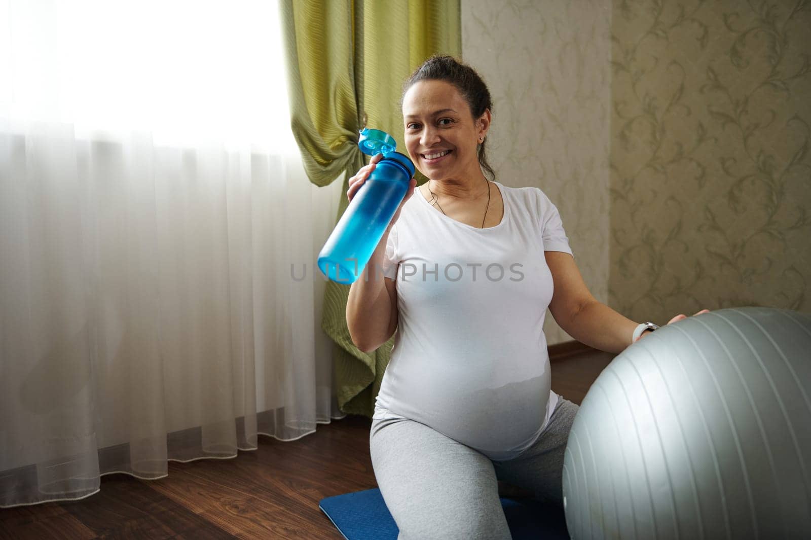 Happy smiling positive cute pregnant woman drinking water from bottle, renewing aqua balance after prenatal fitness workout at home. Healthy active lifestyle during pregnancy. Yoga practice. Maternity