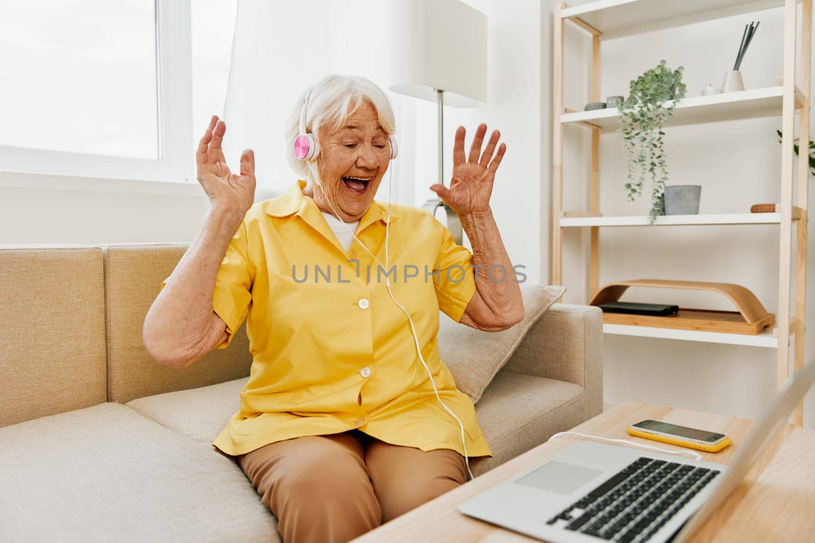 An elderly woman wearing headphones with a laptop communicating online by video call, sitting on the couch at home and working in a yellow shirt in front of a window, the lifestyle of a retired woman. High quality photo