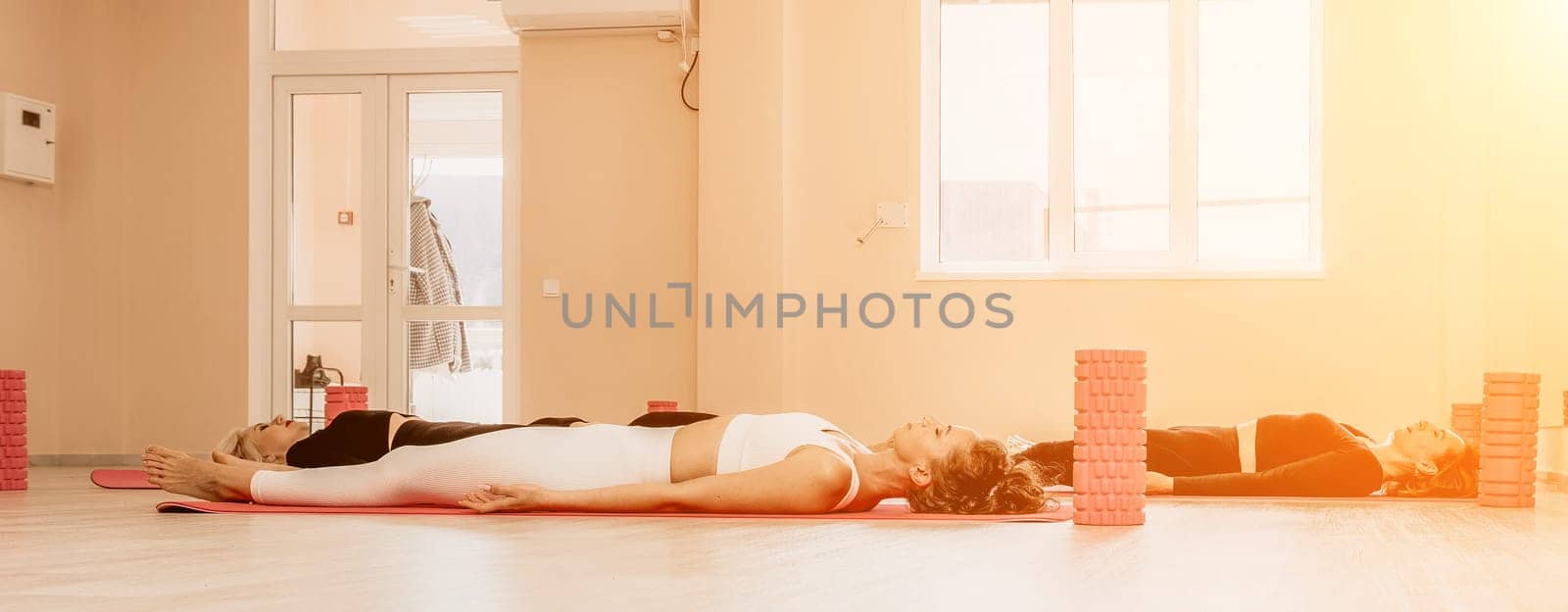 Happy middle aged well looking women, performing fascia exercises on floor using massage foam roller - tool to relieve tension in the back and relieve muscle pain. Female fitness yoga routine concept by panophotograph