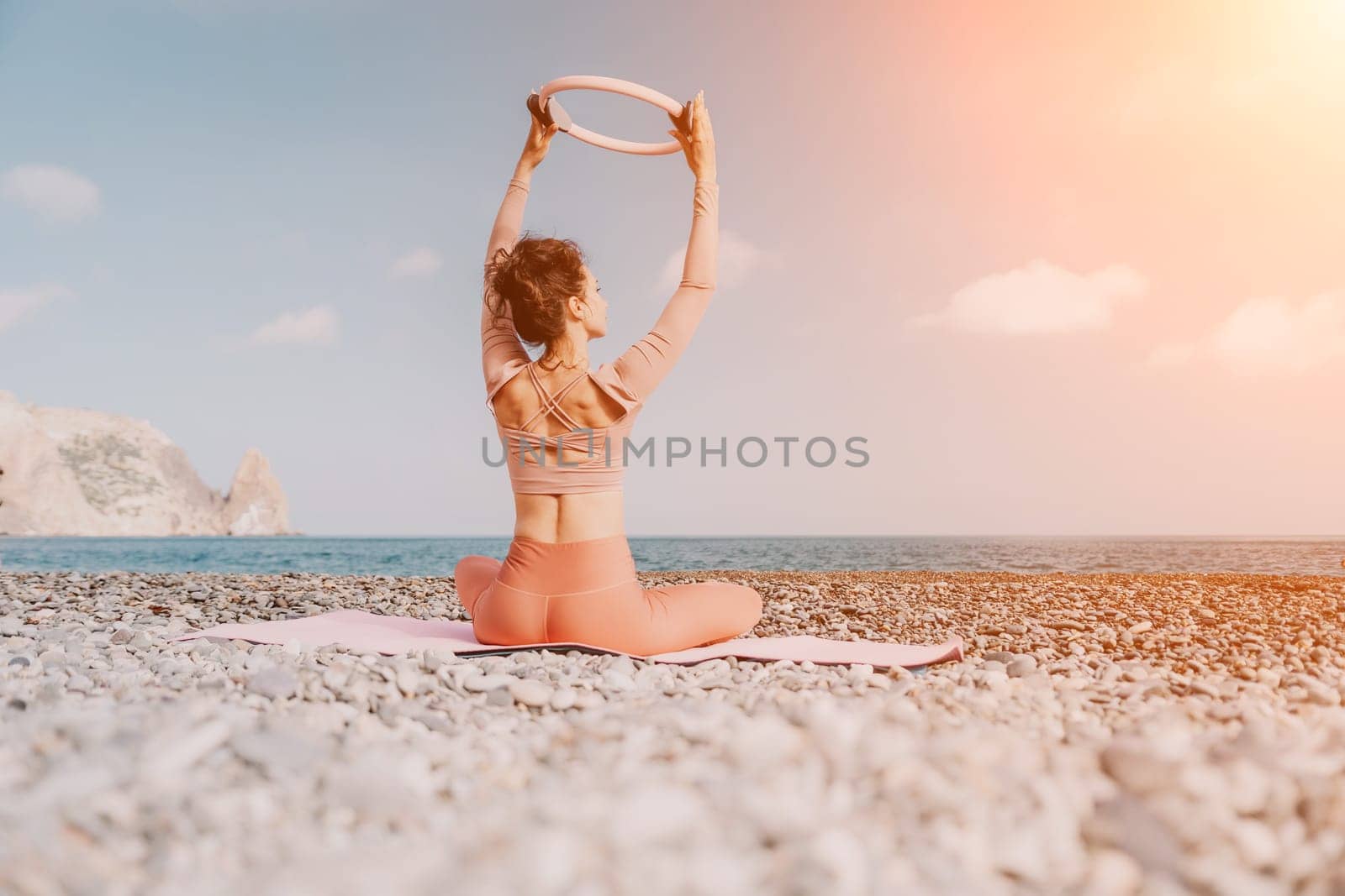 Middle aged well looking woman with black hair doing Pilates with the ring on the yoga mat near the sea on the pebble beach. Female fitness yoga concept. Healthy lifestyle, harmony and meditation.