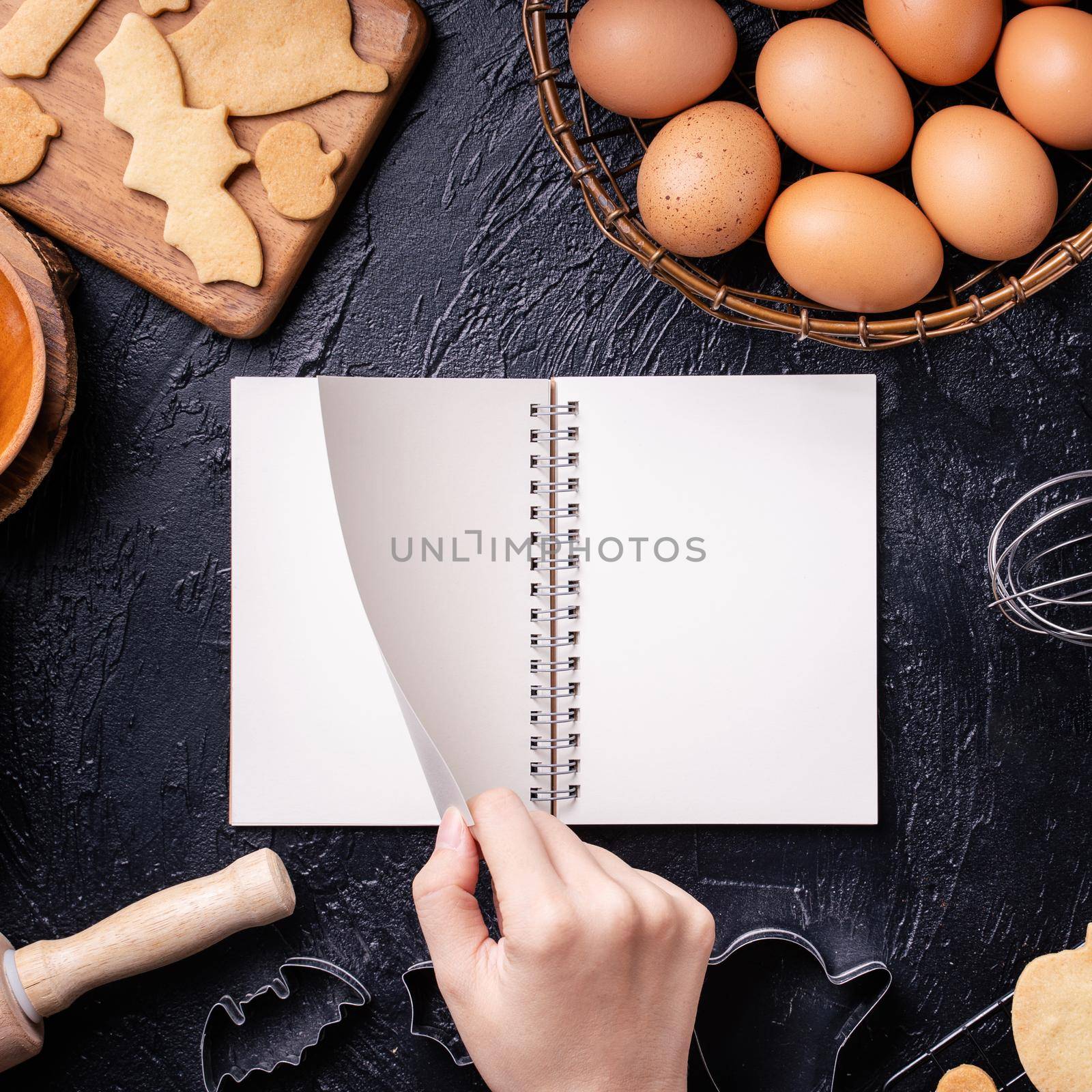 Woman is reading cookbook recipe of making Halloween cookies with baking ingredients, design concept of cooking class, top view, flat lay, overhead.