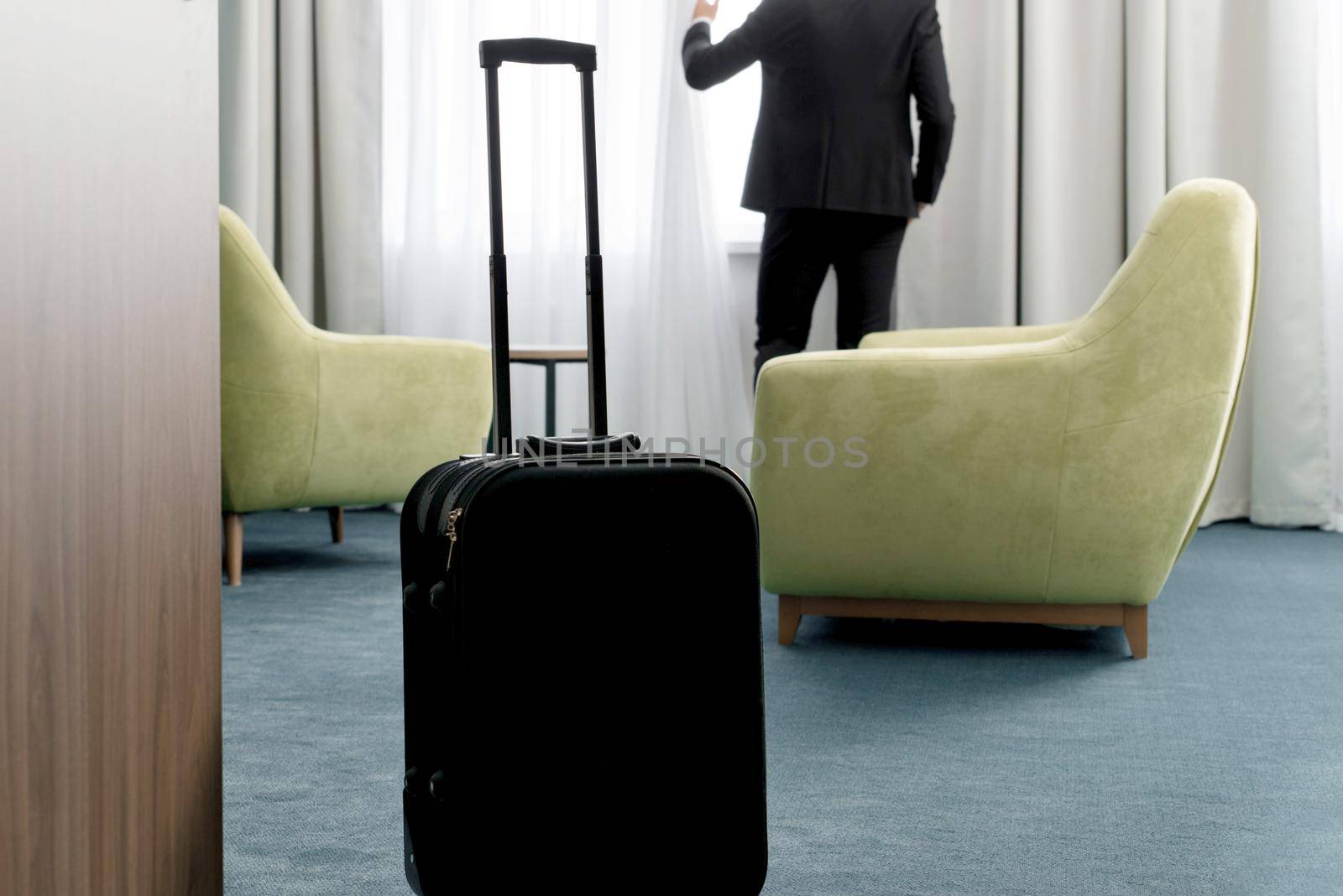 Selective focus of suitcase in hotel room with businessman opening window curtains