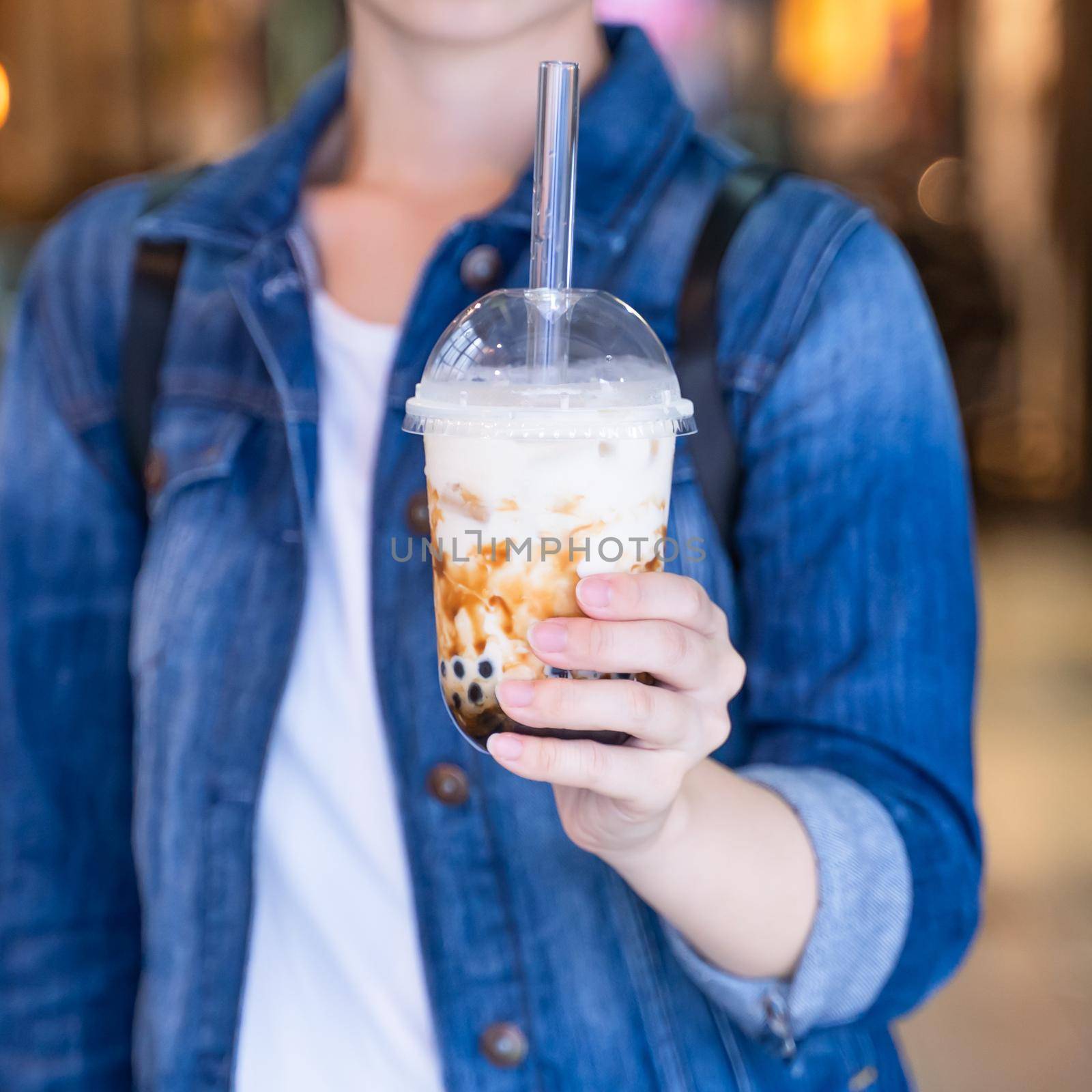 Young girl in denim jacket is drinking brown sugar flavored tapioca pearl bubble milk tea with glass straw in night market of Taiwan, close up, bokeh by ROMIXIMAGE