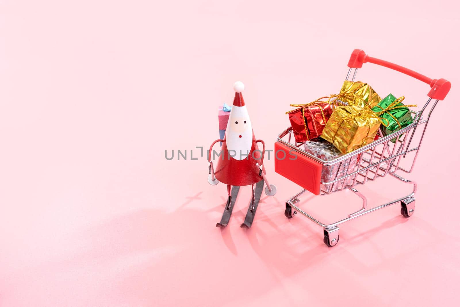 Christmas shopping concept, mini red shop cart trolley with Santa Claus toy and gift box isolated on pale pink background, blank copy space, close up