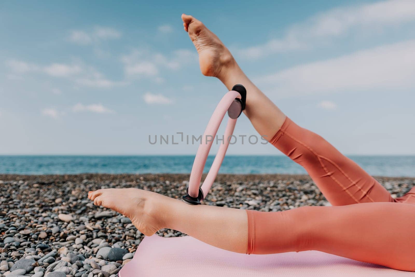 Middle aged well looking woman with black hair doing Pilates with the ring on the yoga mat near the sea on the pebble beach. Female fitness yoga concept. Healthy lifestyle, harmony and meditation.