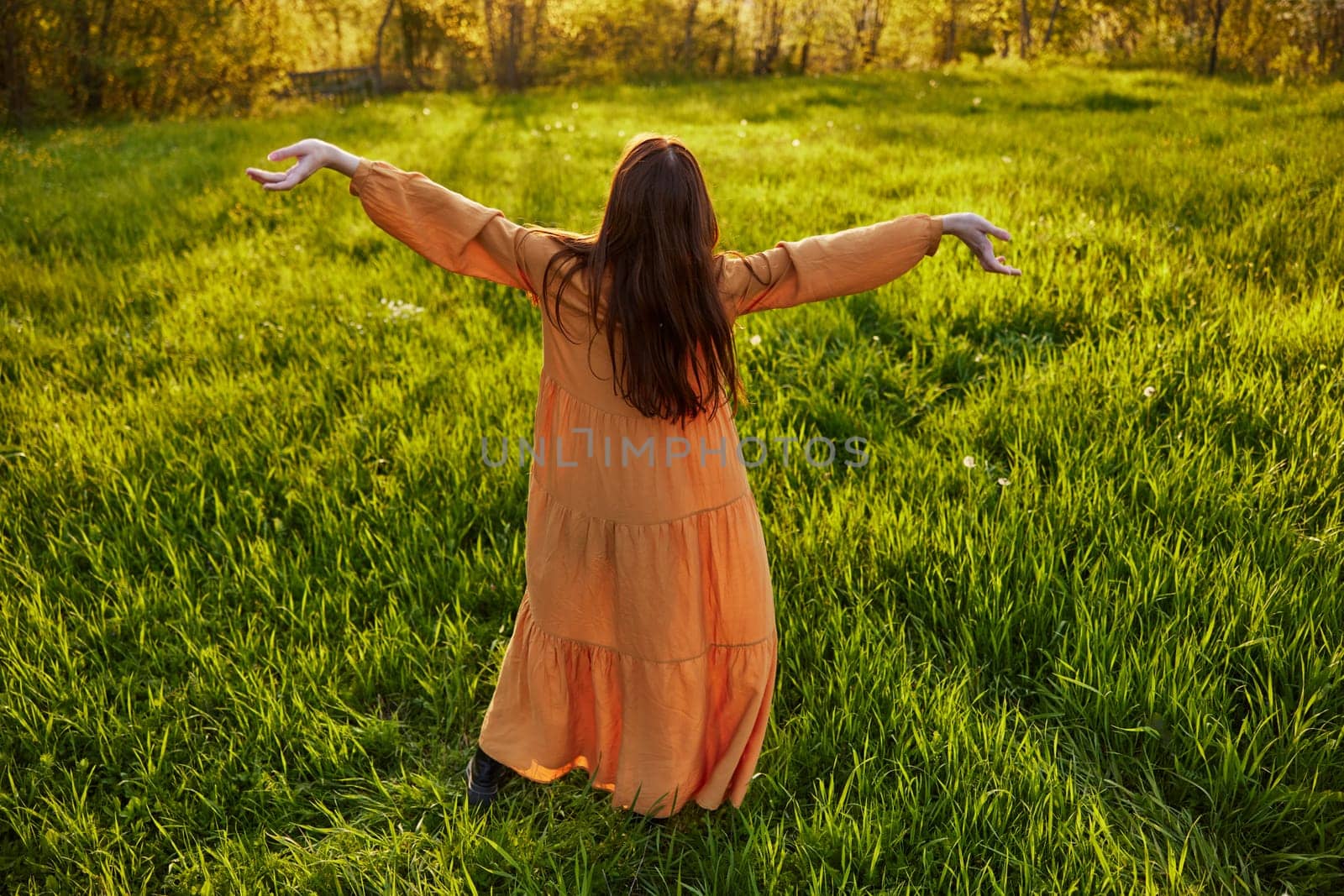 red-haired woman stands in a wide, green field during sunset in a long orange dress enjoying unity with nature and relaxation raising her arms to the sides while standing with her back to the camera by Vichizh