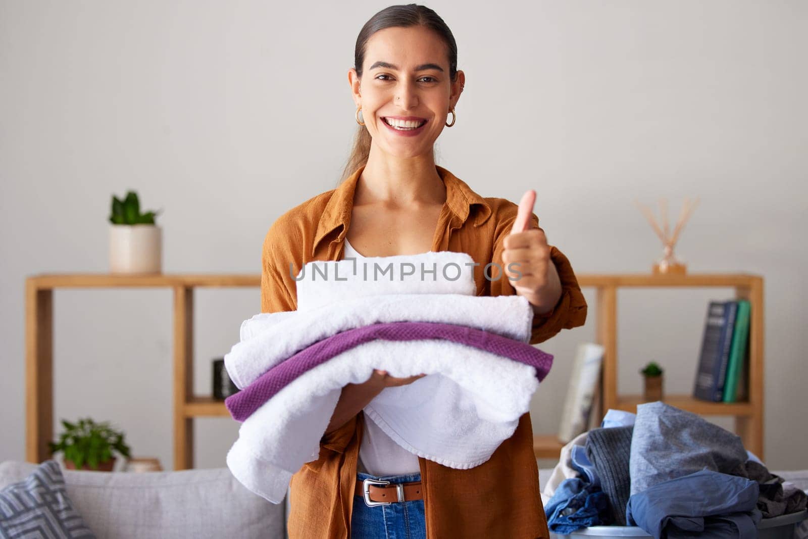 Thumbs up, laundry and portrait of a woman maid folding clothes in the living room in a modern house. Happy, smile and female cleaner or housewife with a thumbsup cleaning or doing chores in a home. by YuriArcurs