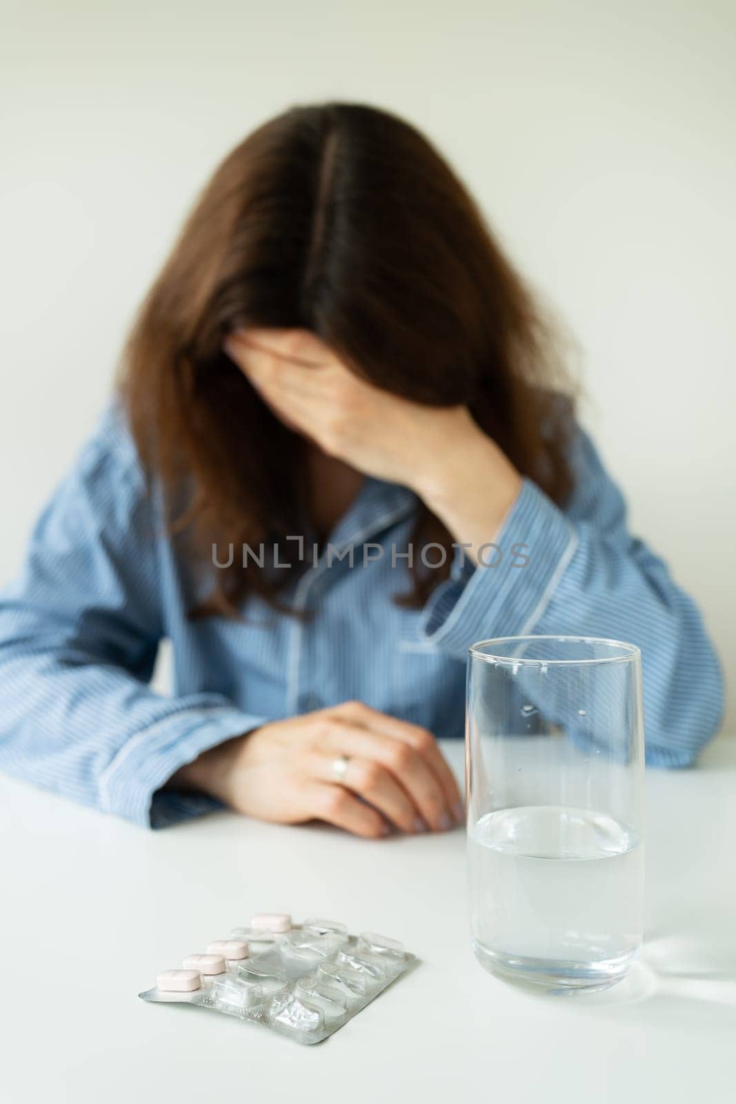 A young girl in pajamas sits at a table and holds her head against the background of a glass of water with pills. The concept of PMS, migraine, stress, depression. by sfinks