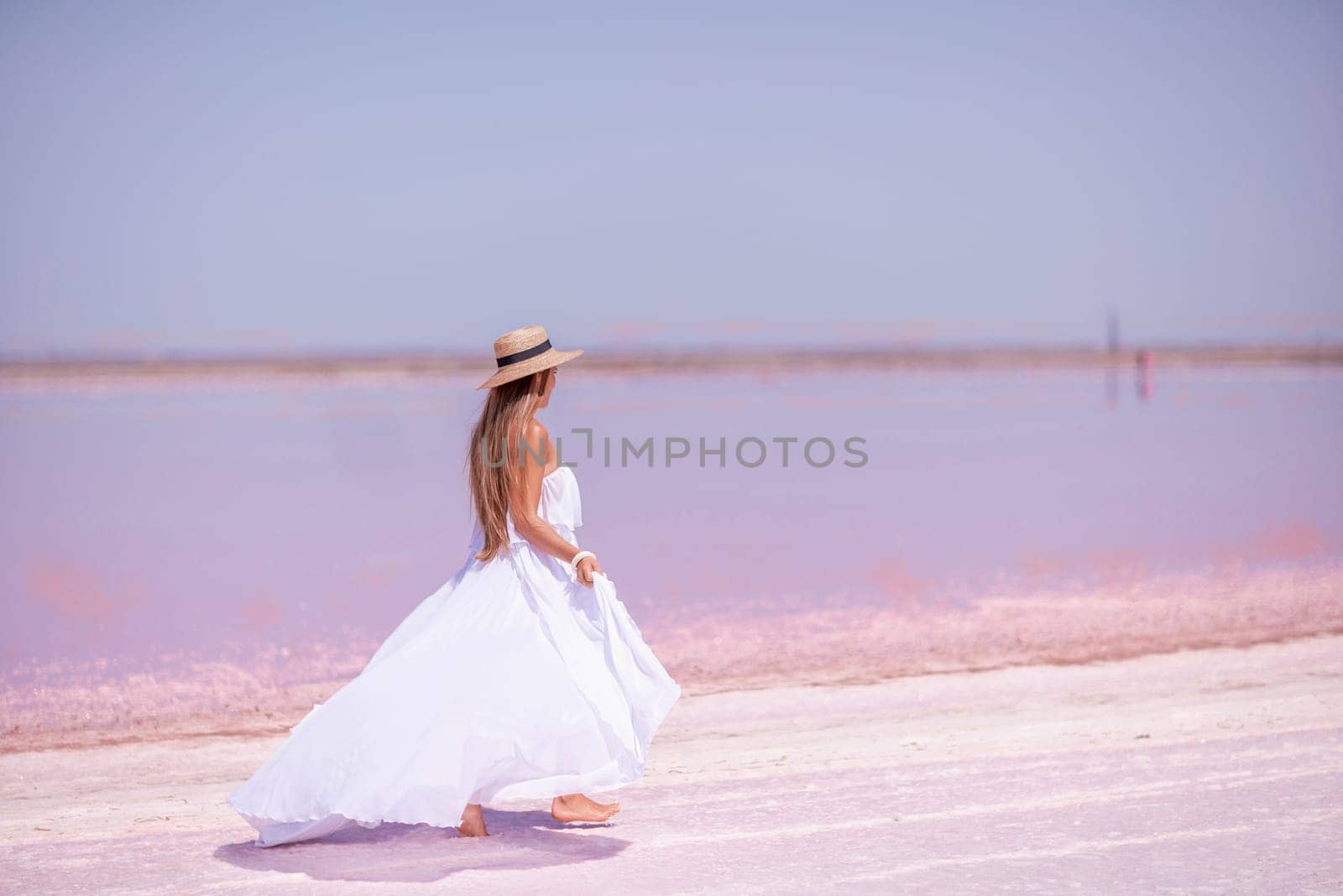 Woman in pink salt lake. She in a white dress and hat enjoys the scenic view of a pink salt lake as she walks along the white, salty shore, creating a lasting memory