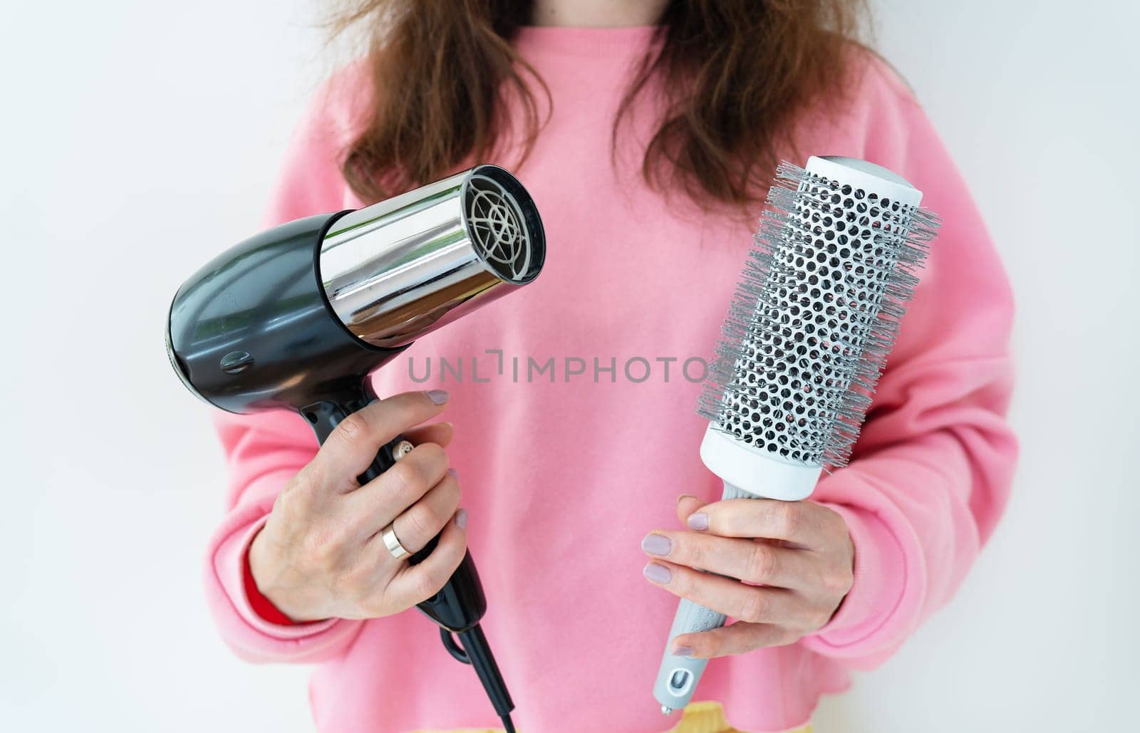 The girl holds a hair dryer and a brush in her hands. Drying long brown hair with a round brush for styling unruly hair. Beauty saloon