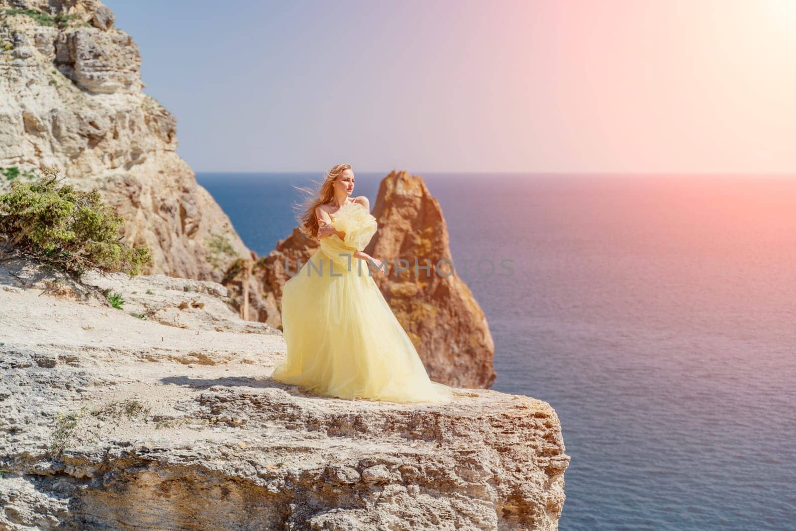 Woman yellow dress sea. Side view Young beautiful sensual woman in yellow long dress posing on a rock high above the sea at sunset. Girl in nature against the blue sky.