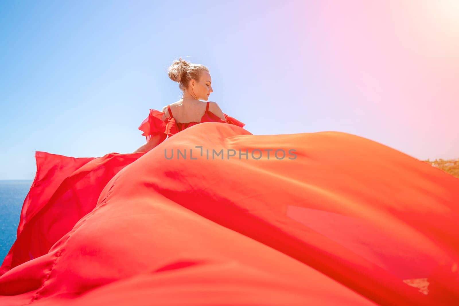 woman sea red dress. Blonde with long hair on a sunny seashore in a red flowing dress, back view, silk fabric waving in the wind. Against the backdrop of the blue sky and mountains on the seashore. by Matiunina