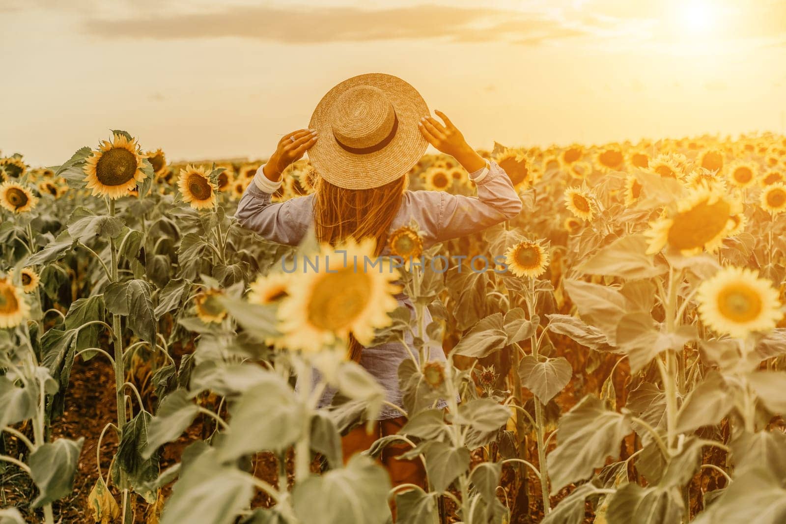 Woman sunflower field. Happy girl in blue dress and straw hat posing in a vast field of sunflowers at sunset. Summer time. by Matiunina