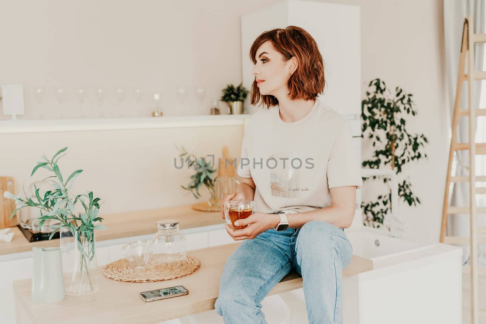 Woman kitchen tea. Carefree woman rests in a cozy kitchen, drinks tea, sits on the table and dreams, free space. Woman holding cup while enjoying hot drink at home by Matiunina