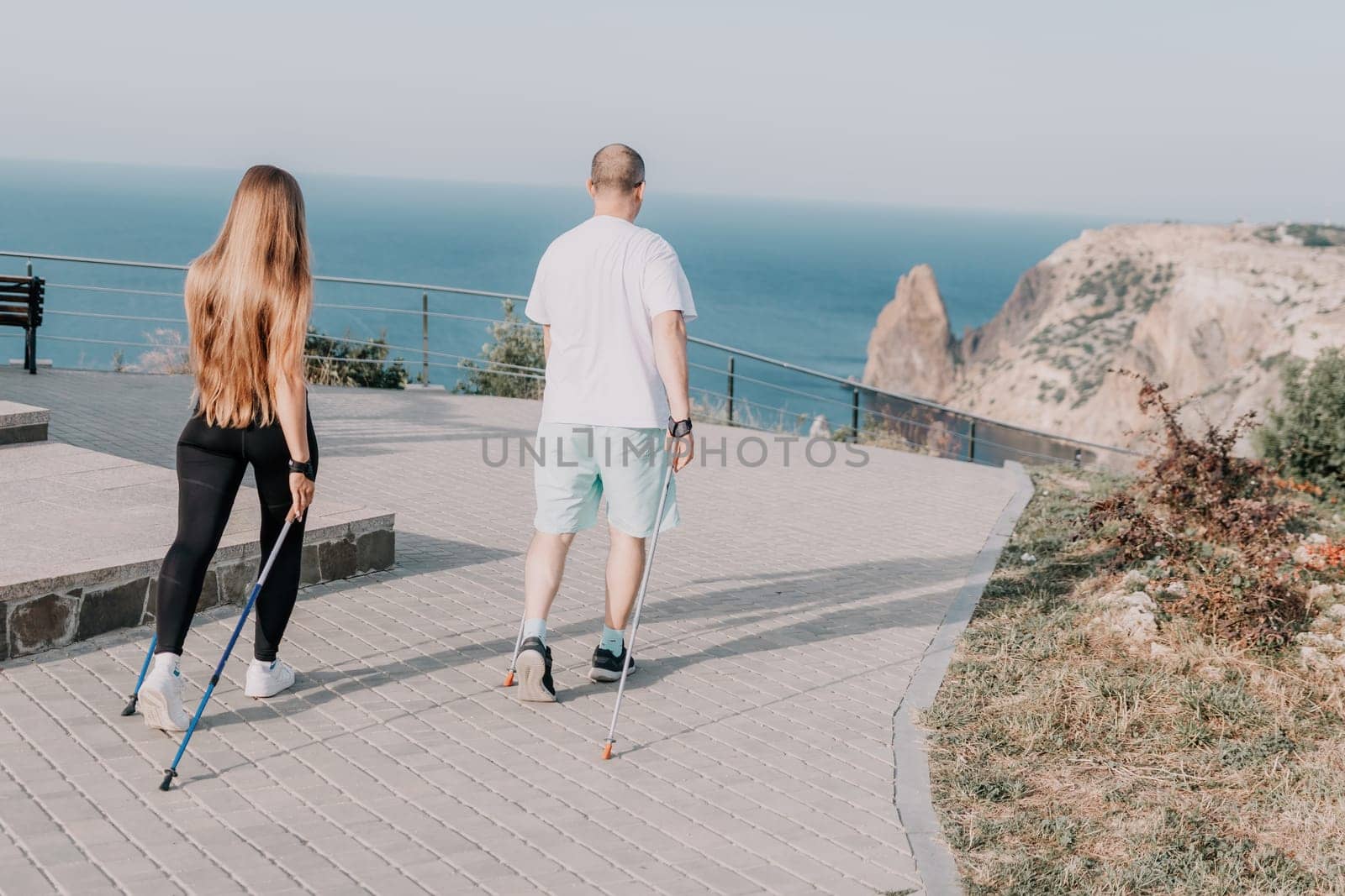 Happy Middle aged couple or friends practicing nordic walking in park near sea. Mature couple with trekking poles walking, practicing Nordic walking outdoors. Aging youthfully and sport concept by panophotograph