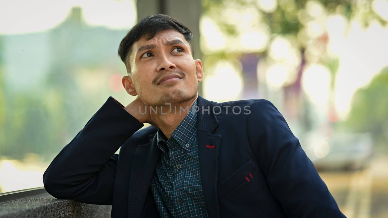 Young businessman in formalwear sitting on bench in urban park and looking thoughtfully.