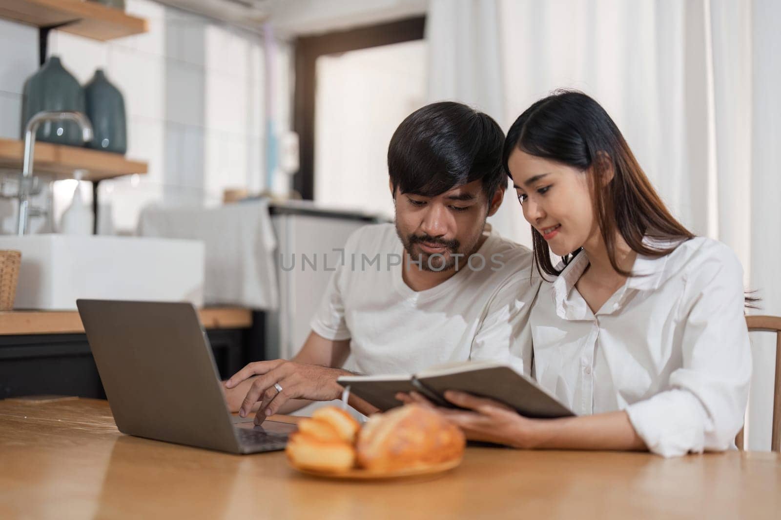 Young asian couple wear casual clothing relaxing while sitting at table in modern kitchen and working with computer laptop at home. Love, happiness, work at home concept by nateemee