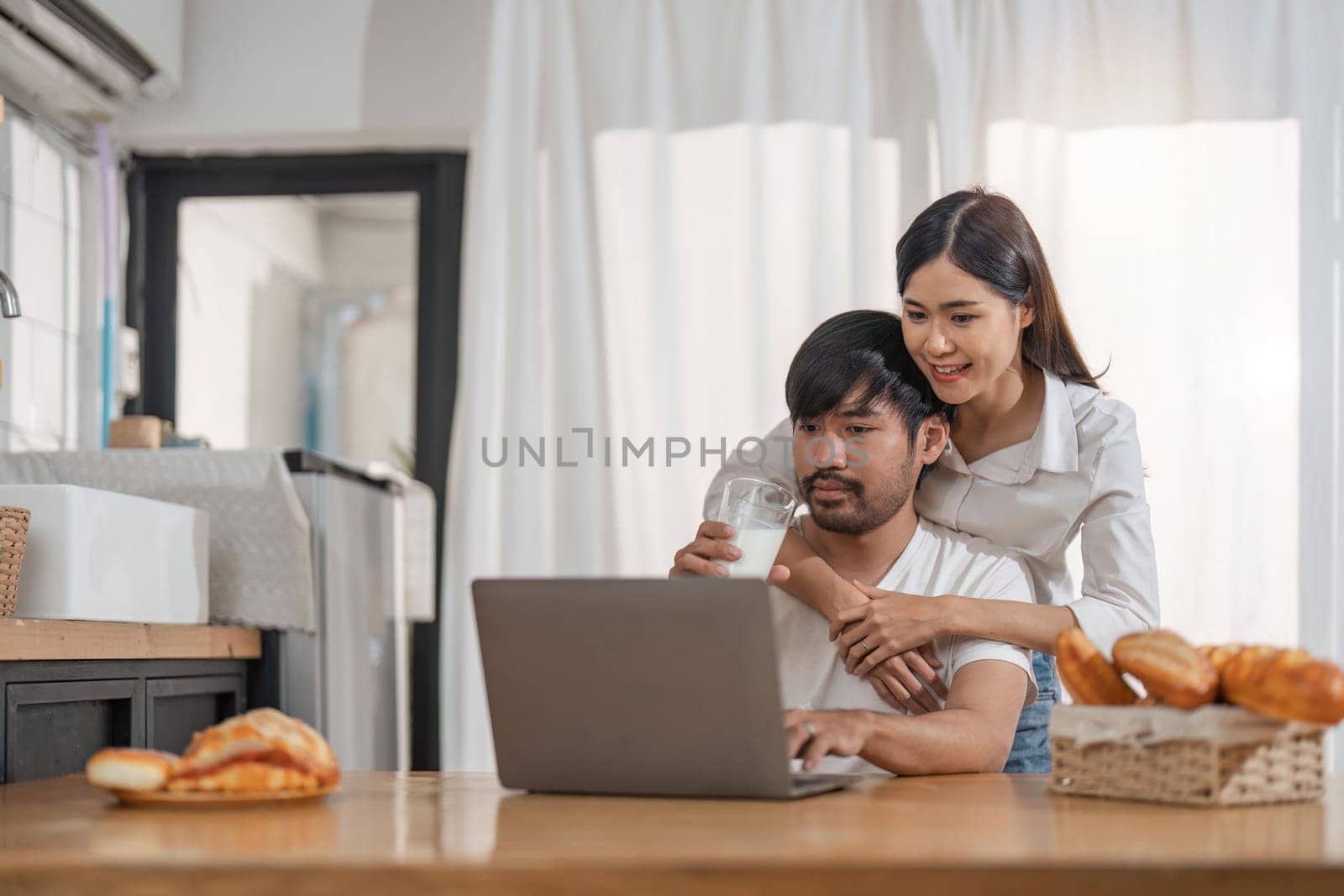 Handsome man sitting near his wife at kitchen. Family couple see social media, surf the web while sitting at kitchen table with generic laptop. Couple working with laptop at home by nateemee