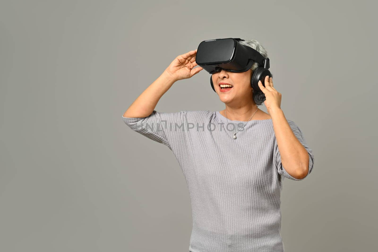 Elderly woman experiencing technology and innovation via VR headset isolated on gray background.