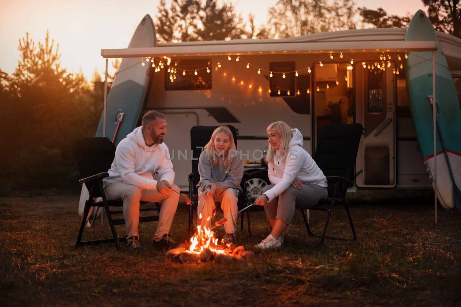 A family cooks sausages on a bonfire near their motorhome in the woods.