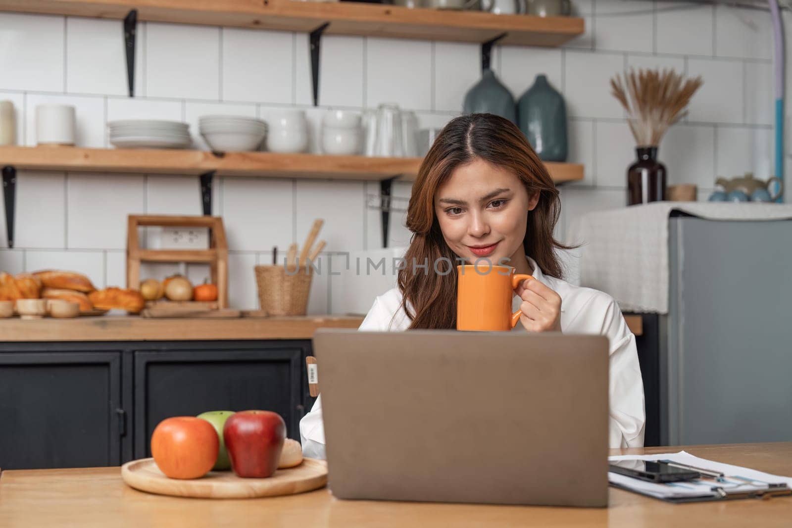 Young woman drinking morning coffee and looking at monitor of laptop while sitting in the kitchen at home.