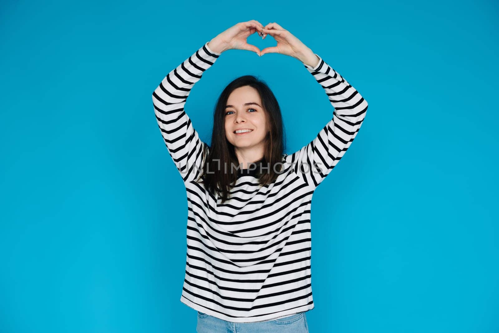 Joyful Woman in Striped Sweater Expressing Love and Happiness - Happy Girl Showing Heart Gesture with Arms, Smiling - Isolated Blue Background with Copy Space - Perfect for Love, Happiness Concepts