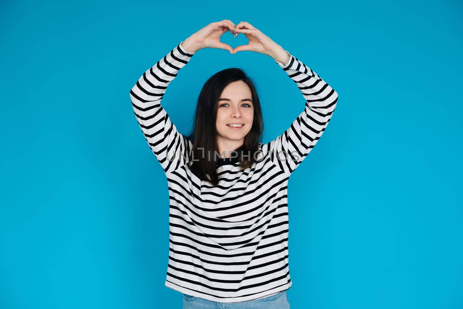 Happy Woman in Striped Sweater Spreading Love and Positivity - Smiling Girl with Raised Arms Forming Heart Shape, Expressing Joy and Affection - Isolated Blue Background - Ideal for Happiness, Love,