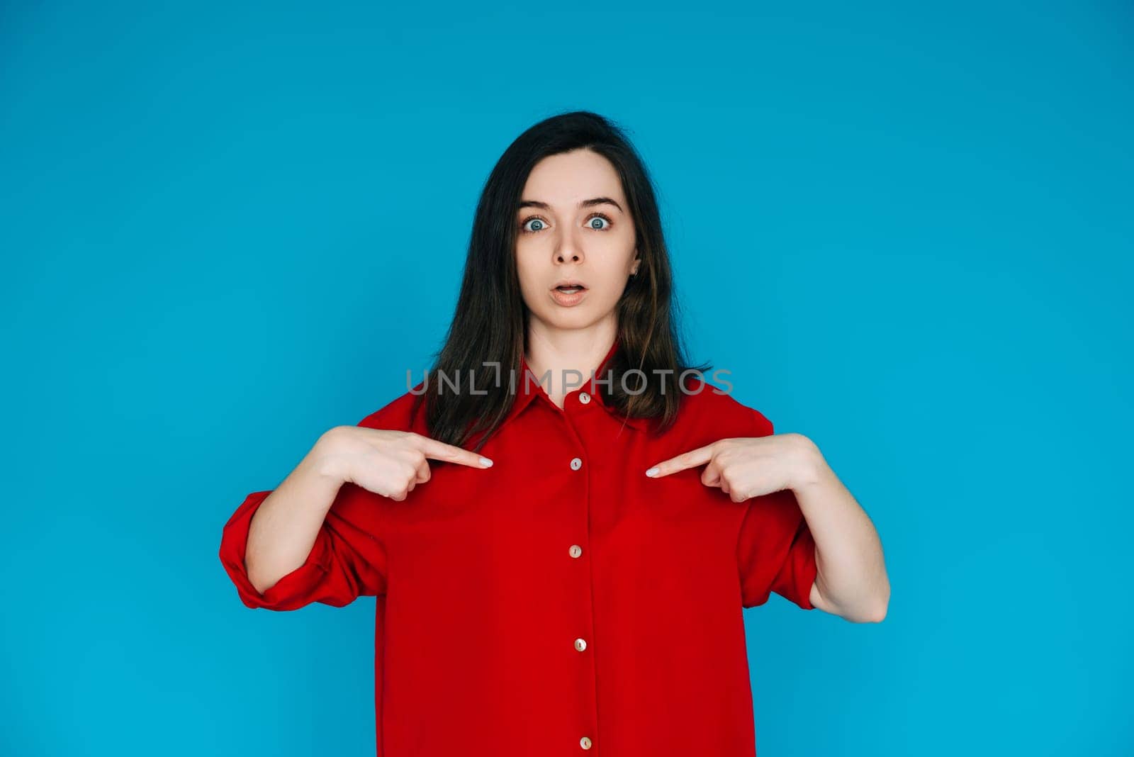 A photo of a lovely impressed young woman in a red shirt, surprised face, pointing her finger, isolated on a blue background - Perfect for Emphasizing Surprise and Amazement.