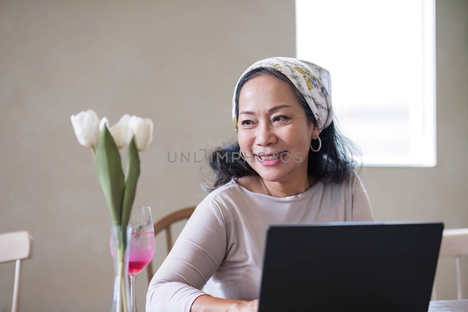 Attractive beautiful mature Asian woman sits at a table with her use laptop in her minimal living room. Hobby and lifestyle concepts by wichayada