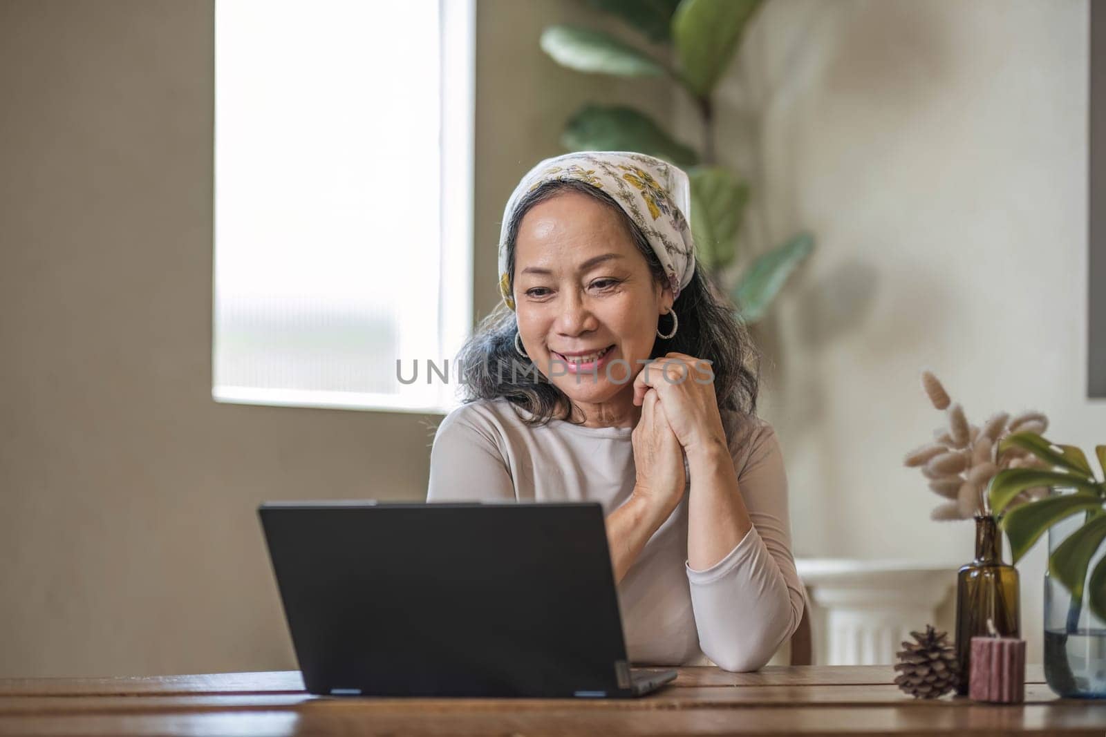 Attractive beautiful mature Asian woman sits at a table with her use laptop in her minimal living room. Hobby and lifestyle concepts by wichayada