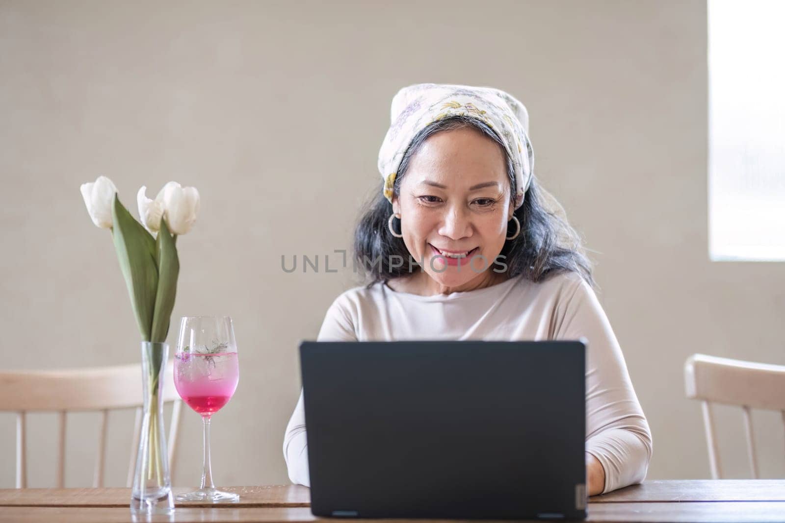 Attractive beautiful mature Asian woman sits at a table with her use laptop in her minimal living room. Hobby and lifestyle concepts by wichayada