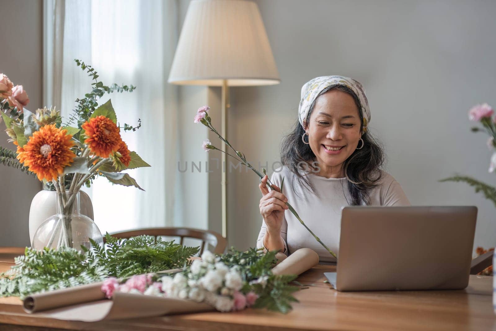 Happy and attractive mature Asian woman enjoying with her online flower arrangement workshop at home. by wichayada